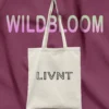 Natural cotton canvas tote bag with LIVNT logo, hanging on a rich plum fabric background under pastel "WILDBLOOM" text