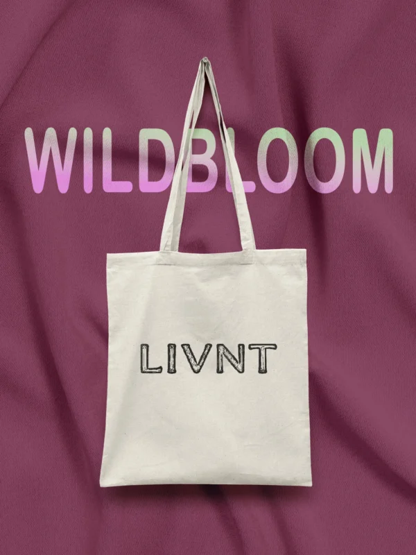 Natural cotton canvas tote bag with LIVNT logo, hanging on a rich plum fabric background under pastel "WILDBLOOM" text