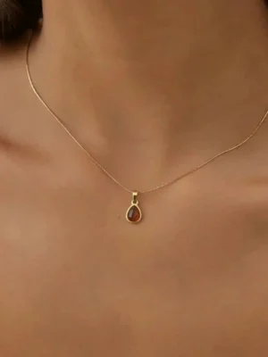 A woman wearing the LIVNT Amber Drop Necklace, which features a delicate gold chain and a small, pear-shaped amber-colored stone pendant, resting elegantly on her collarbone against a soft white top.