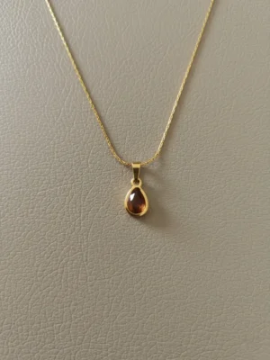 The LIVNT Amber Drop Necklace, featuring a delicate gold chain and a small, pear-shaped amber-colored stone pendant, meticulously photographed on a minimalist, premium textured grey-beige leather background with subtle shadows.