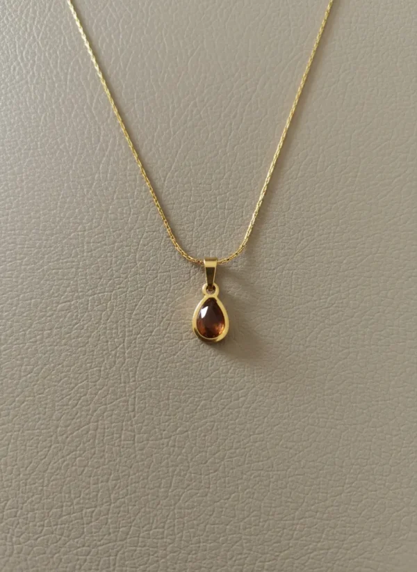 The LIVNT Amber Drop Necklace, featuring a delicate gold chain and a small, pear-shaped amber-colored stone pendant, meticulously photographed on a minimalist, premium textured grey-beige leather background with subtle shadows.