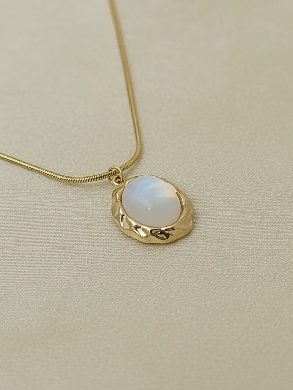 Close-up of the Pebble Anti-Tarnish Necklace showing the iridescent stone and textured gold setting resting on a soft cream surface.