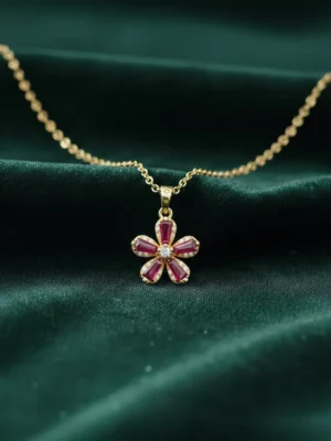 A gold Scarlet Flower pendant necklace with five ruby-red baguette-cut gemstone petals and a central diamond, resting on a dark green silk background.