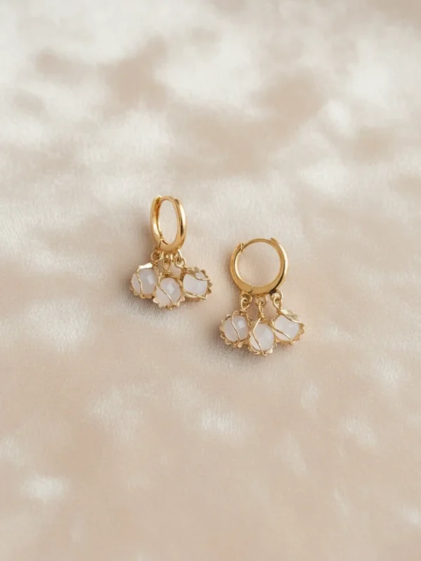 Aura Whisper Hoops in 18K gold with white gemstone clusters, set on a soft, light gold satin fabric. The image emphasizes the warmth of the gold and the ethereal quality of the white stones.