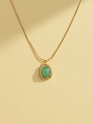 A high-quality product shot of a gold pendant necklace on a solid cream-colored background. The necklace features a smooth snake chain and a cushion-cut, light green stone in a gold setting with a twisted rope border, with a soft shadow to the side.