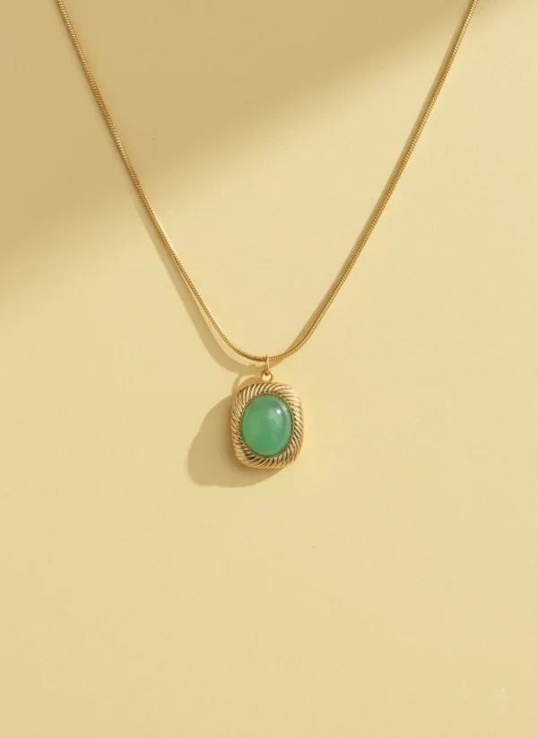 A high-quality product shot of a gold pendant necklace on a solid cream-colored background. The necklace features a smooth snake chain and a cushion-cut, light green stone in a gold setting with a twisted rope border, with a soft shadow to the side.
