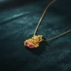 Close-up of a matte gold Temple Lakshmi pendant with two deep magenta ruby-toned Kemp stones, suspended on a fine micro-chain, elegantly resting on rich, dark teal satin fabric.