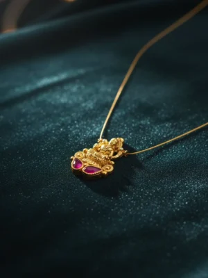 Close-up of a matte gold Temple Lakshmi pendant with two deep magenta ruby-toned Kemp stones, suspended on a fine micro-chain, elegantly resting on rich, dark teal satin fabric.