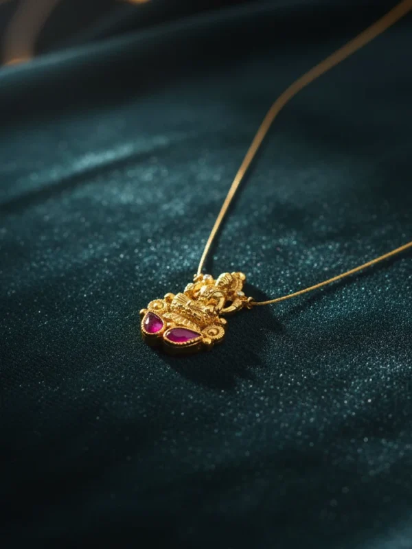 Close-up of a matte gold Temple Lakshmi pendant with two deep magenta ruby-toned Kemp stones, suspended on a fine micro-chain, elegantly resting on rich, dark teal satin fabric.