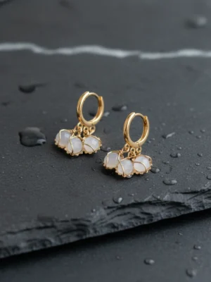 Detailed close-up of Aura Whisper Moonstone Gold Hoops against a dark, textured slate background with water droplets. Captures the contrast and brilliance of the 18K gold and white gem clusters.