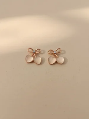 A pair of Cupid's Bow Earrings in rose gold with opalescent heart charms, styled on a neutral beige background with soft, elegant lighting.