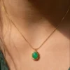 A close-up of a woman's neck and collarbone, showcasing a delicate gold snake chain necklace with a square green gemstone pendant. The vibrant green stone is set in a gold bezel with a detailed twisted rope border, catching the warm sunlight.