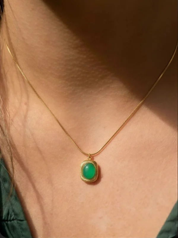 A close-up of a woman's neck and collarbone, showcasing a delicate gold snake chain necklace with a square green gemstone pendant. The vibrant green stone is set in a gold bezel with a detailed twisted rope border, catching the warm sunlight.