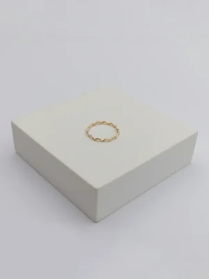 A close-up, angled product shot of the gold Wave Ring. The focus is on the ring's delicate, rippling silhouette as it sits on the corner of a white display block.